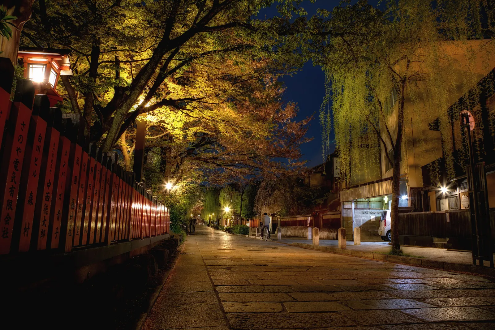 A narrow stone street in Gion at night, lit by lanterns, with willow trees and wooden buildings along the canal