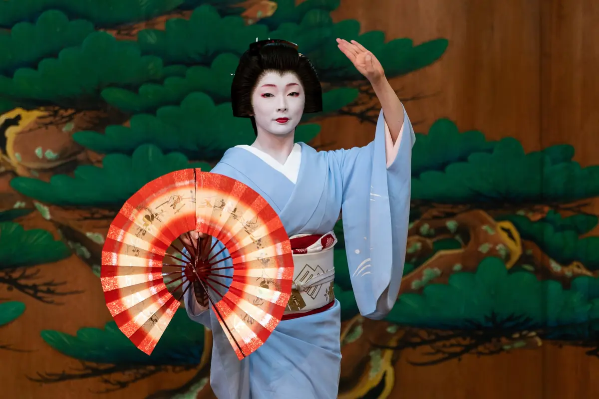 A geiko in a pale blue kimono stands on a stage in front of a painted pine screen, holding two red and white folding fans open to her sides