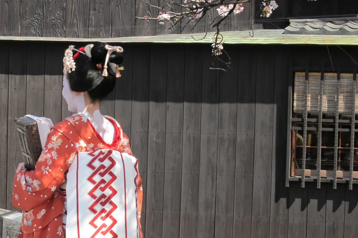 A woman dressed as a maiko, seen from behind in an orange floral kimono and a long white obi, beside a wooden bridge rail in Gion