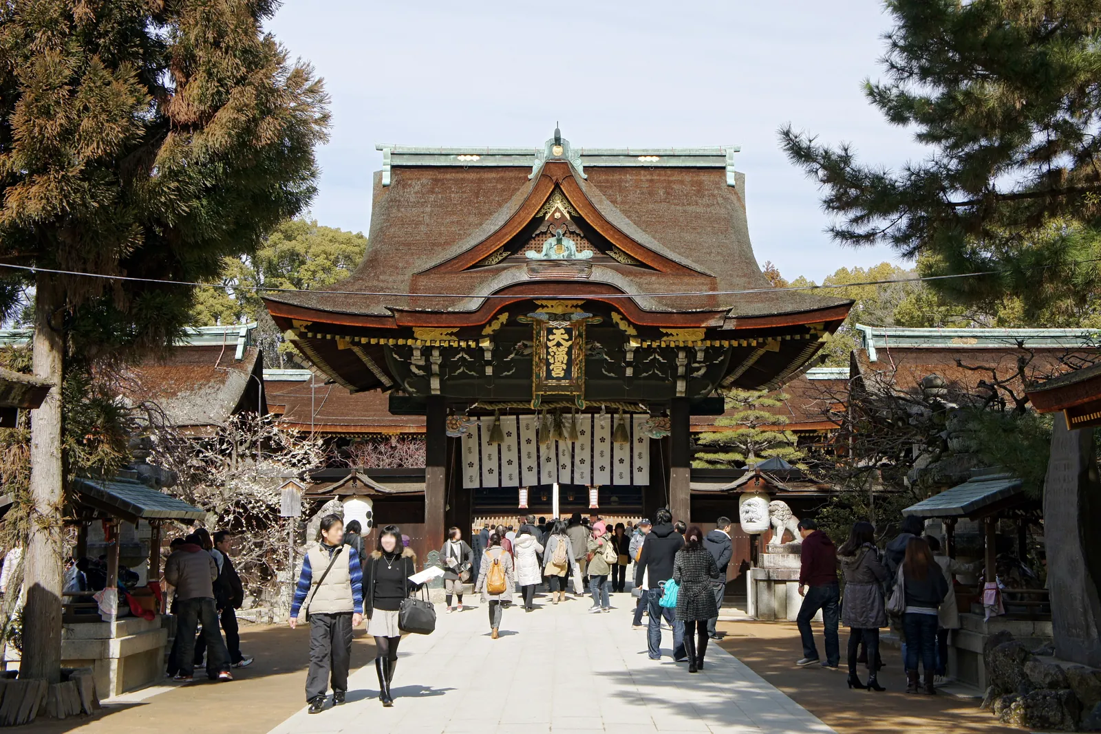 The main gate of Kitano Tenmangū in Kyoto, with plum trees in blossom and visitors walking up the stone approach under a blue sky